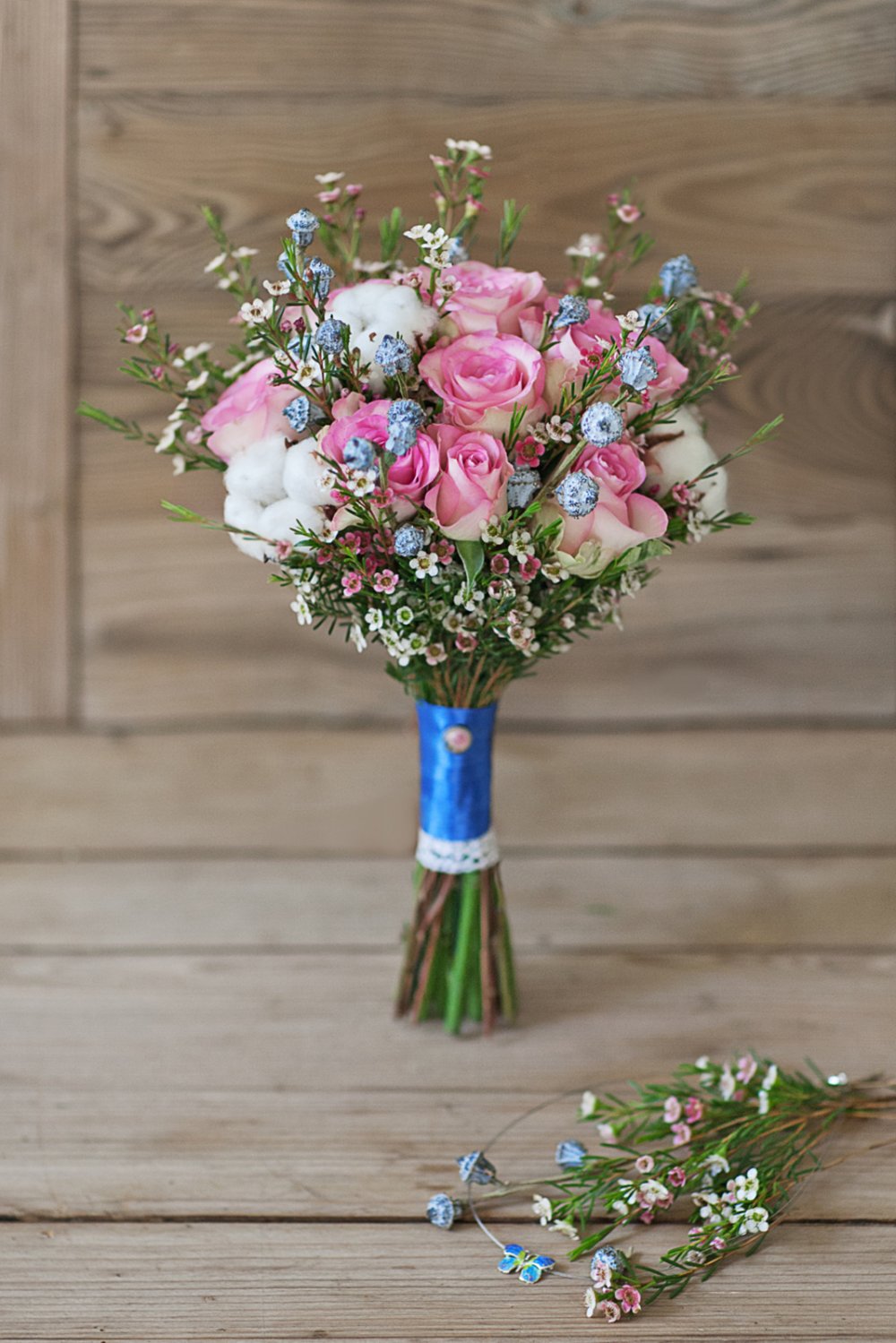 Розово-голубой свадебный букет из роз, ваксфлауэра, коробочек натурального хлопка и голубых ягод. Украшен кружевом, атласной лентой и декоративной брошью. Букет идеально подходит для зимней свадьбы, он стойкий и не боится низких температур. 